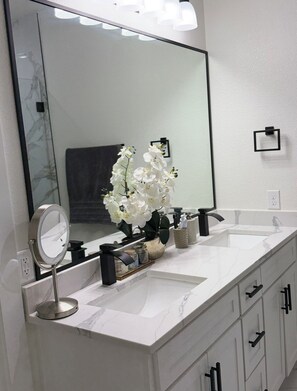 A bathroom with a large mirror, white marble countertop, a sink, and a vase with white flowers. - 3-bedroom house in charming Fort Worth with AC for relaxing stays (Fort Worth)