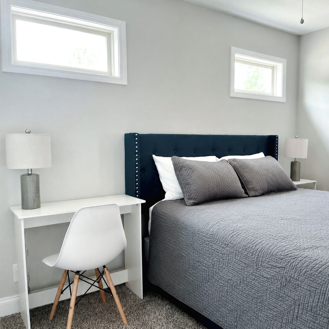A bedroom with a bed, two bedside tables, a desk, a chair, and two windows.