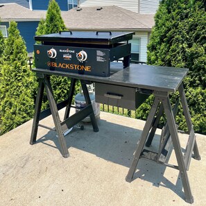 En portabel grill med reglage och en gastank, placerad utomhus med ett hus och träd i bakgrunden.