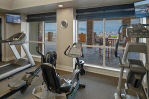 Een sportschool met loopbanden, een elliptische trainer en een hometrainer, met uitzicht op een haven.