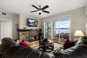A living room with a fireplace, leather sofas, a television, and a balcony with a view. - Lakefront Luxury Downtown w/ Pool, Hot Tub & Dock (Sandpoint)