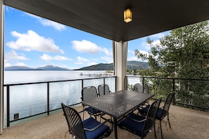 A terrace with a table and chairs, overlooking a calm body of water and distant mountains. - Lakefront Luxury Downtown w/ Pool, Hot Tub & Dock (Sandpoint)