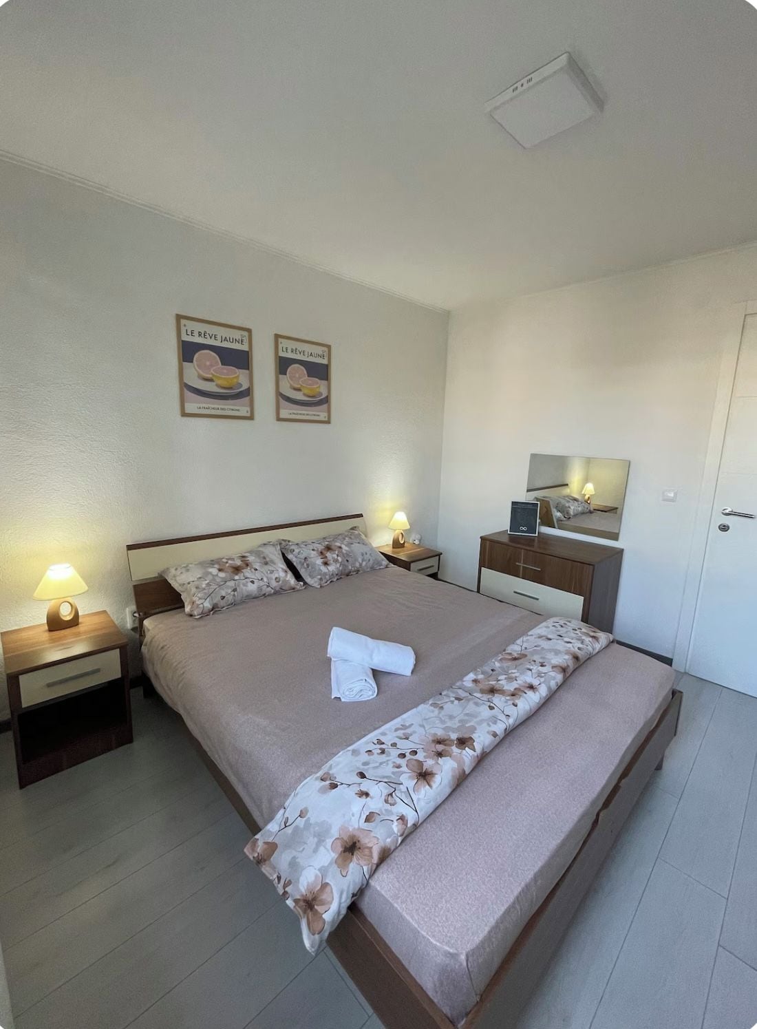 Una cama bien tendida con una colcha estampada, dos lámparas de noche, una mesita de noche, un aparador y un espejo en la pared.