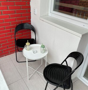 Un pequeño espacio al aire libre con una mesa blanca y dos sillas negras, una pared de ladrillo rojo y una ventana con persianas.