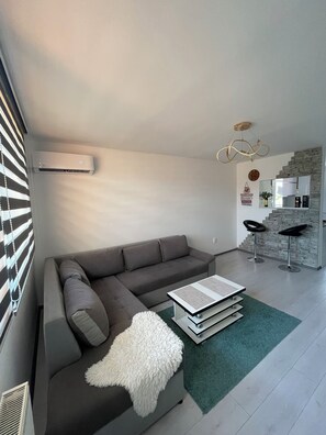 Una sala de estar moderna con un sofá seccional gris, un plaid blanco suave, una mesa de centro y una cocina con salpicadero de piedra y dos taburetes de bar.