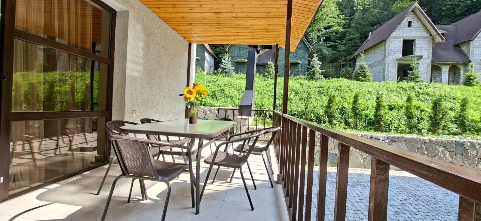Un balcón con mesa y sillas, una pérgola de madera y vistas a una casa y vegetación.