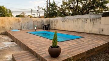 A rectangular swimming pool with a wooden deck surrounding it, a potted plant, and a small table with chairs.