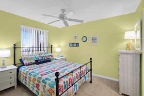 A bedroom with a bed, nightstands, a ceiling fan, a window with blinds, a wall clock, and a small table with a lamp. - Atlantic Sunrise-Oceanfront-Minutes from Flagler-2Bed/2Ba (New Smyrna Beach)