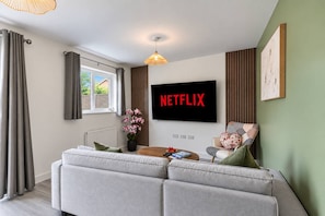 A modern living room with a sofa, a coffee table, a flat-screen TV, and a window with curtains. - Spacious 4BR Home | Sleeps 8 | Parking + Garden (Quedgeley)