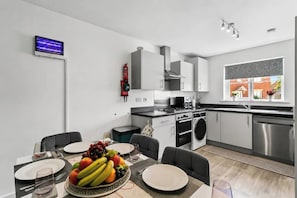 A modern kitchen with a dining area, a fire extinguisher, and a window with a view of buildings. - Spacious 4BR Home | Sleeps 8 | Parking + Garden (Quedgeley)