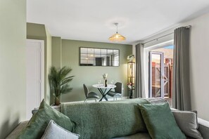 A room with a green sofa, a dining table with chairs, a potted plant, and a large window with curtains. - Spacious 4BR Home | Sleeps 8 | Parking + Garden (Quedgeley)