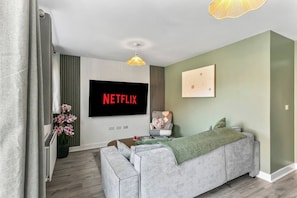 A modern living room with a grey sofa, a flat-screen TV displaying 'NETFLIX', a decorative chair, and a wall-mounted light fixture. - Spacious 4BR Home | Sleeps 8 | Parking + Garden (Quedgeley)