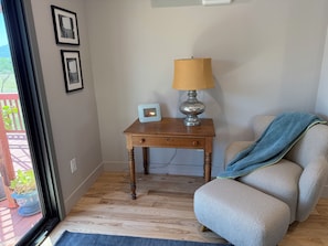 Een kamer met een houten vloer, een klein tafeltje met een lamp, een lijst met een afbeelding en een stoel met een plaid.