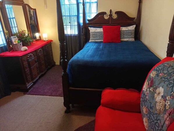 A bedroom with a wooden bed, a red pillow, a floral armchair, and a dresser with a mirror.