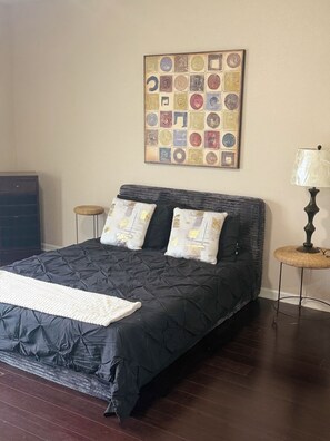 A bedroom with a black quilted bedspread, two pillows with a gold pattern, a white throw blanket, a wall-mounted quilted artwork, a wooden nightstand with a lamp, and a dark wooden dresser. - Awesome 3-bedroom house with AC in quaint Denham Springs (Denham Springs)