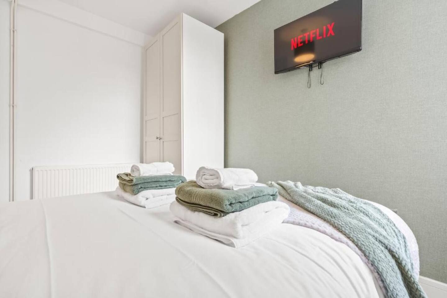 Una camera d'albergo con un letto, una televisione a muro che mostra 'NETFLIX' e una pila di asciugamani piegati.