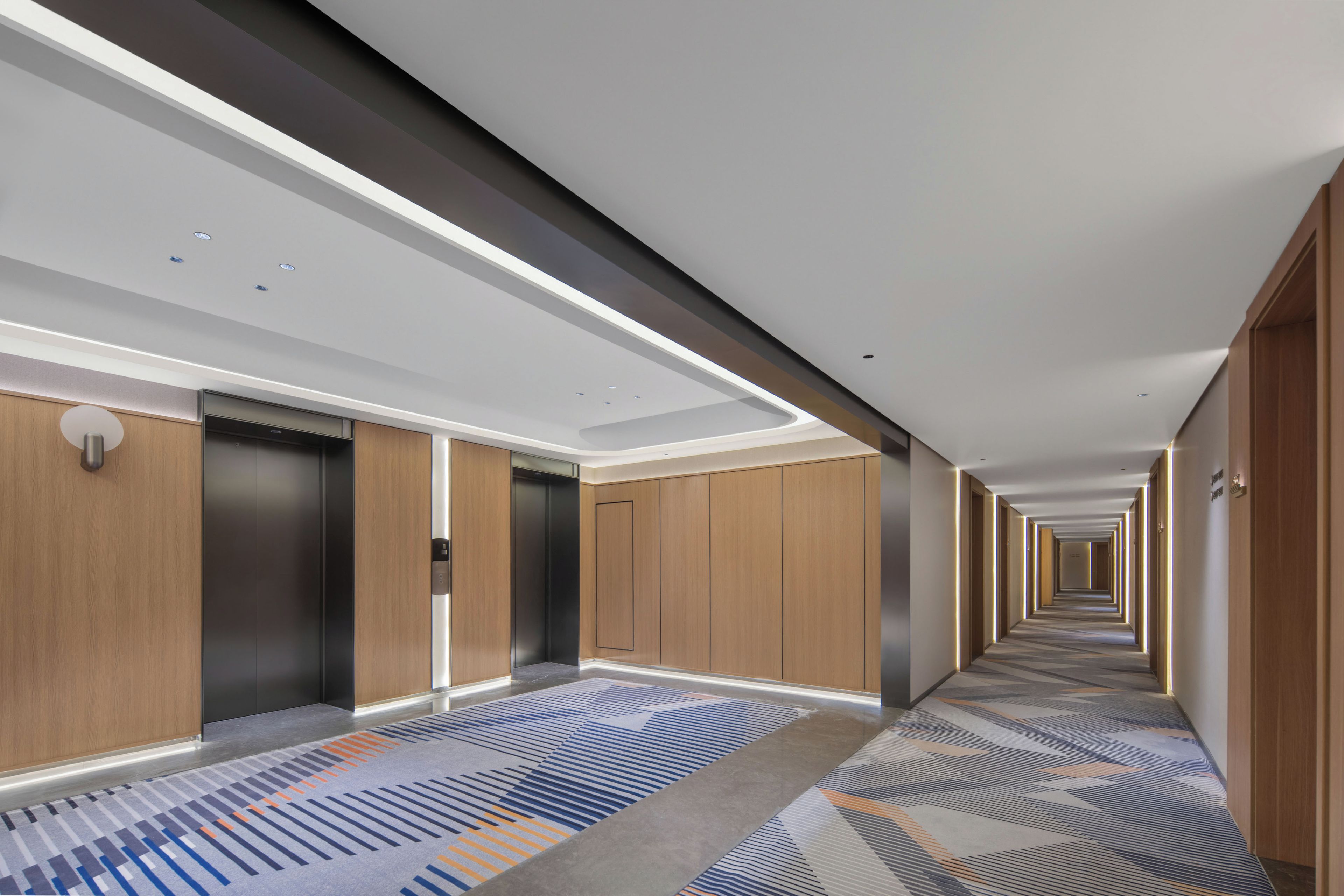 A modern hotel corridor with a patterned carpet, wooden panel walls, and a long hallway.