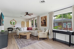 A modern living room with a sofa, coffee table, and chairs, a dining area with a table and chairs, and a large window with a view of greenery. - Sanford Pool and Patio Retreat (Sanford)
