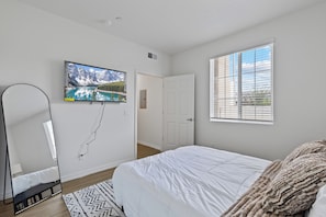 Ein Schlafzimmer mit einem Bett, einem an der Wand montierten Fernseher, einem Fenster mit Jalousien und einer Tür.