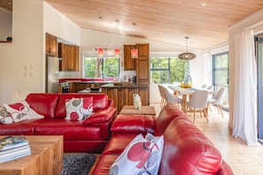 A modern living room with a red leather sofa, wooden dining table, and a kitchen area with wooden cabinets and a refrigerator. - The Tasting Room by Waiheke Pure, close to wineries, great valley views (Auckland)