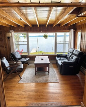 A cabin interior with a wooden floor, a sofa, a table, and a large window overlooking a lake. - Lakefront Maine Getaway - Private Dock & Kayaks (Burnham)