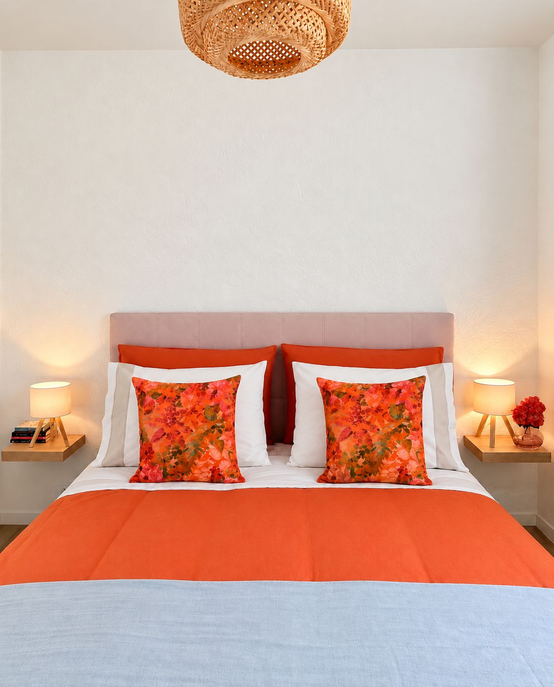 A bed with an orange bedspread, two white pillows, and two orange floral pillows. There is a woven headboard, two bedside tables with lamps, and a wall-mounted light fixture.