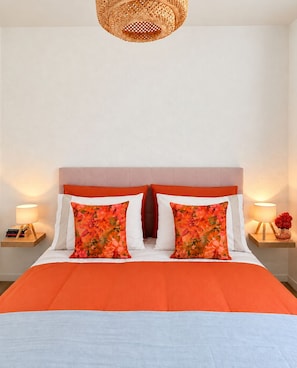 A bed with an orange bedspread, two white pillows, and two orange floral pillows. There is a woven headboard, two bedside tables with lamps, and a wall-mounted light fixture. - Fiumicino Downtown Vacation Rental with Self-Check-In (Fiumicino)
