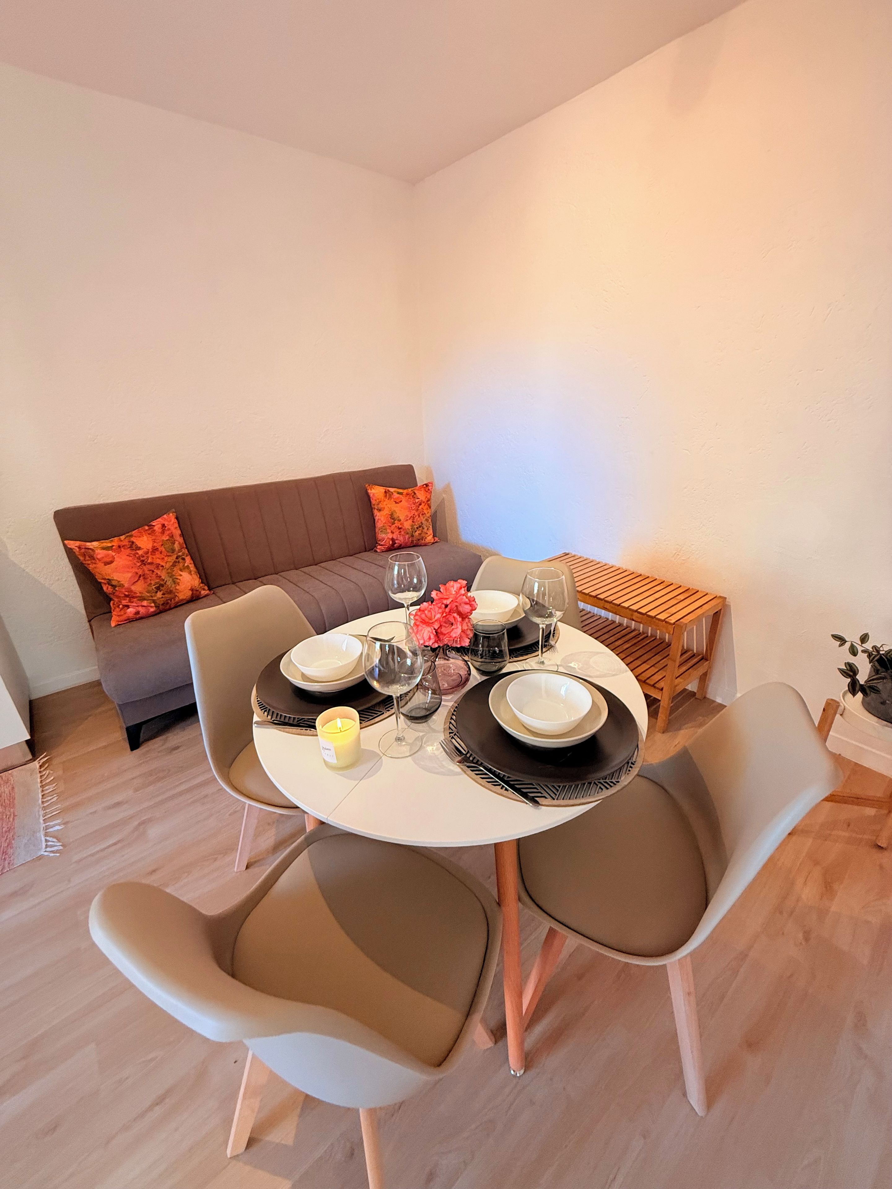 A dining area with a round table set for four, surrounded by chairs. A sofa with cushions is positioned against the wall.