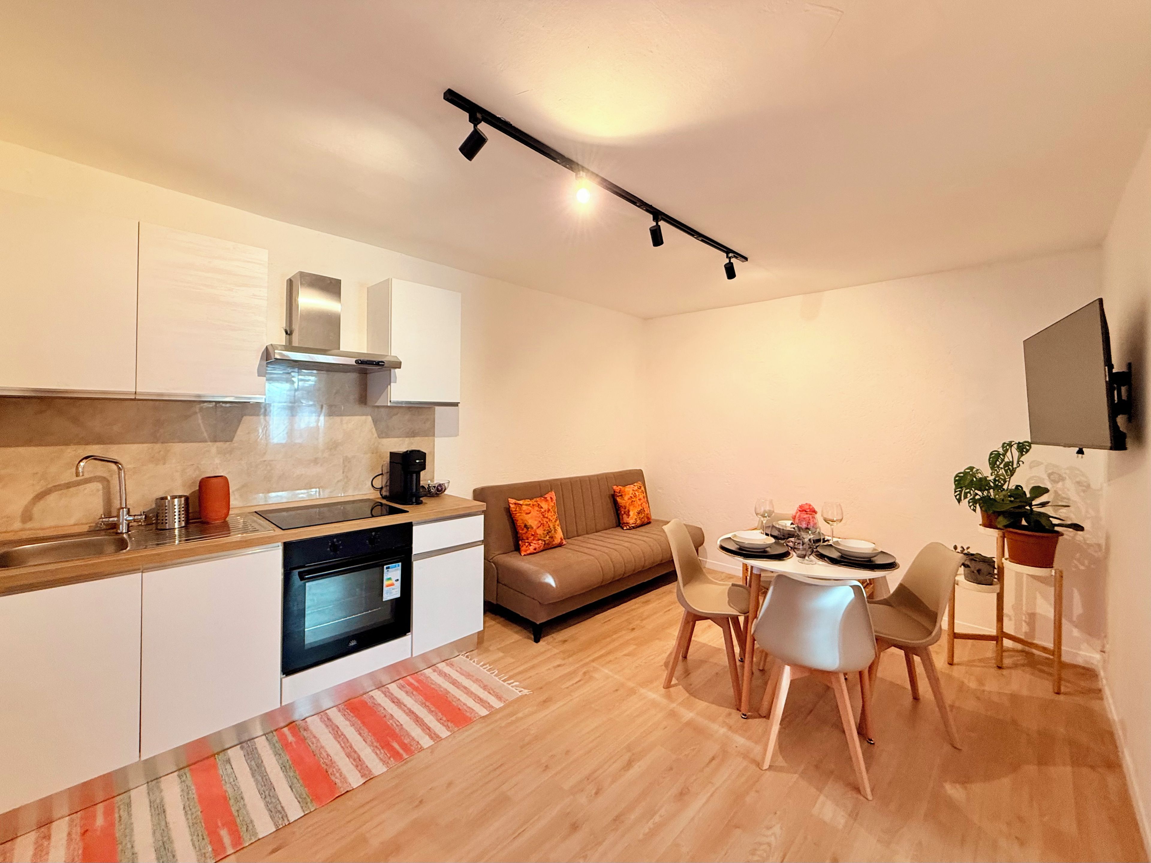 A modern kitchen with a built-in oven, a dining area with a table set for four, and a sofa with orange cushions.