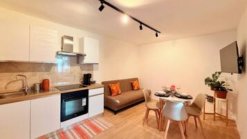 A modern kitchen with a built-in oven, a dining area with a table set for four, and a sofa with orange cushions.
