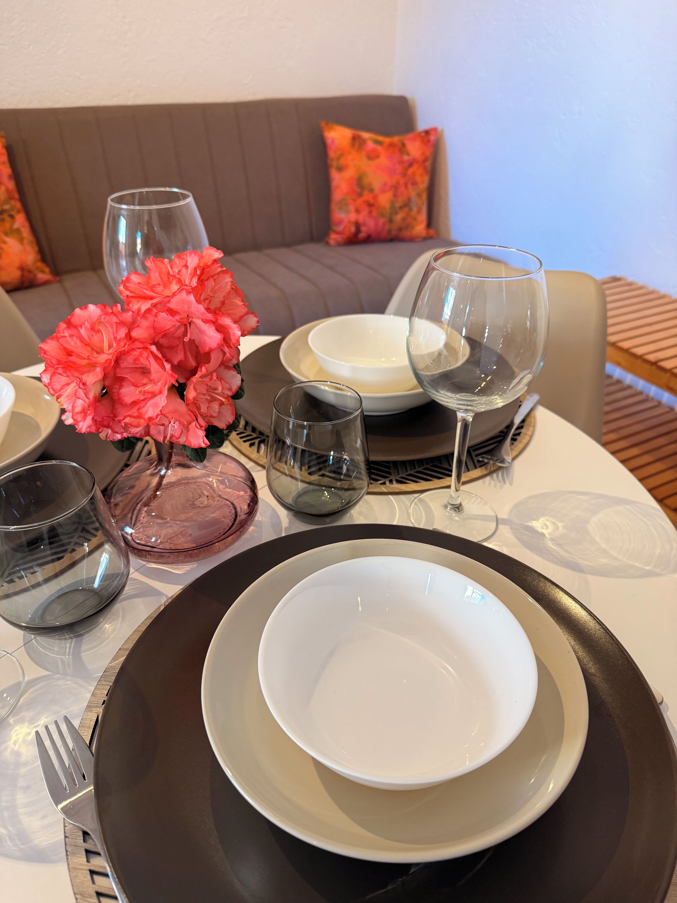 A dining table set with plates, glasses, and cutlery, with a sofa and decorative pillows in the background.