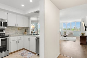 A modern kitchen with stainless steel appliances and white cabinetry, a dining area with a sofa and table, and a view of greenery through the windows. - Endless Summer! Salty vibes only. (St. Petersburg)