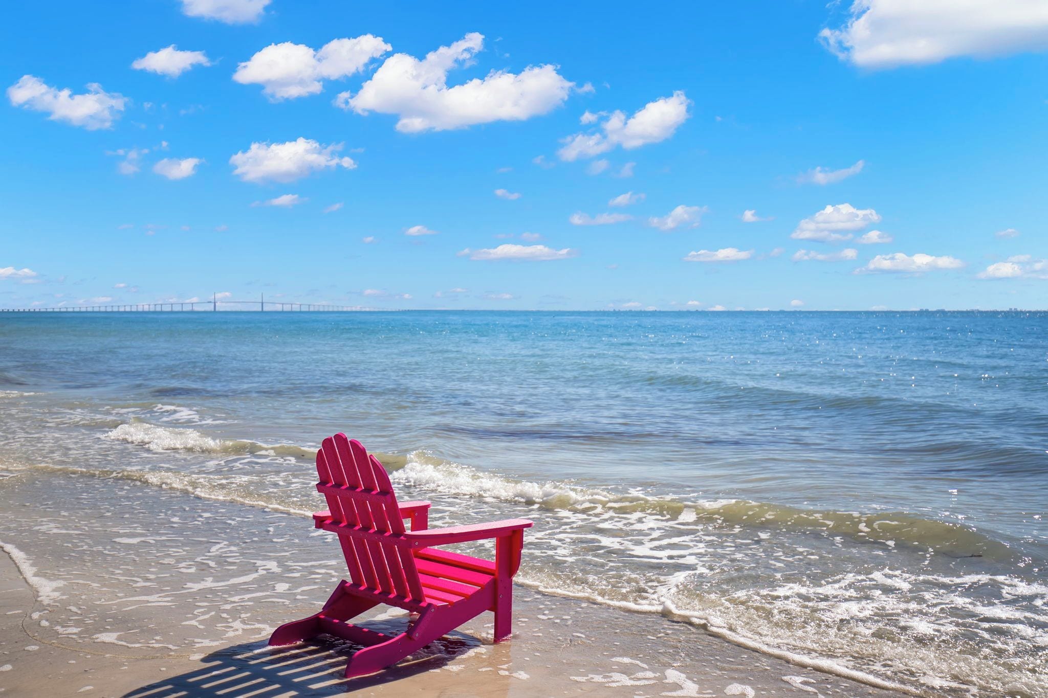 A beach scene with a pink Adirondack chair on the sand, a clear blue sky, and a bridge in the distance.