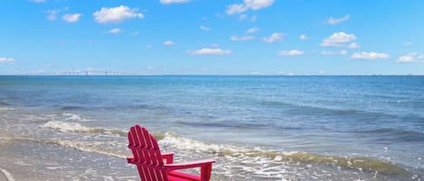 Escena de playa con una silla Adirondack rosa sobre la arena, cielo azul despejado y un puente a lo lejos.