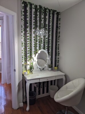 Una habitación con un tocador, un espejo con luces, una silla y una pared decorada con enredaderas verdes y un letrero.
