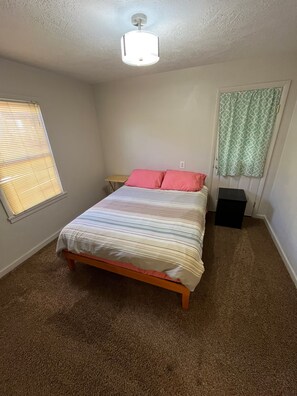 Een slaapkamer met een bed, een nachtkastje, een raam met jaloezieën, een plafondlamp en een deur met een decoratief gordijn.