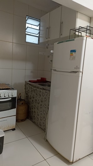 A kitchen with white cabinets, a gas stove, a refrigerator, and a window with blinds. - Apartamento na Barra (Salvador)