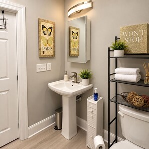 A bathroom with a white sink, a mirror, a toilet, and a shelf with towels and a plant. - Nascar King Suite - 12 Mins to Speedway (Kannapolis)