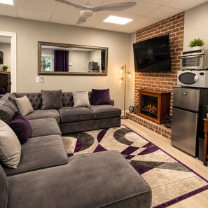 A living room with a sectional sofa, a fireplace, a flat-screen TV, and a modern kitchen area with a microwave and oven. - Nascar King Suite - 12 Mins to Speedway (Kannapolis)