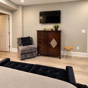 A living room with a grey sofa, a wooden cabinet, a television mounted on the wall, and a small wooden stool. - Nascar King Suite - 12 Mins to Speedway (Kannapolis)