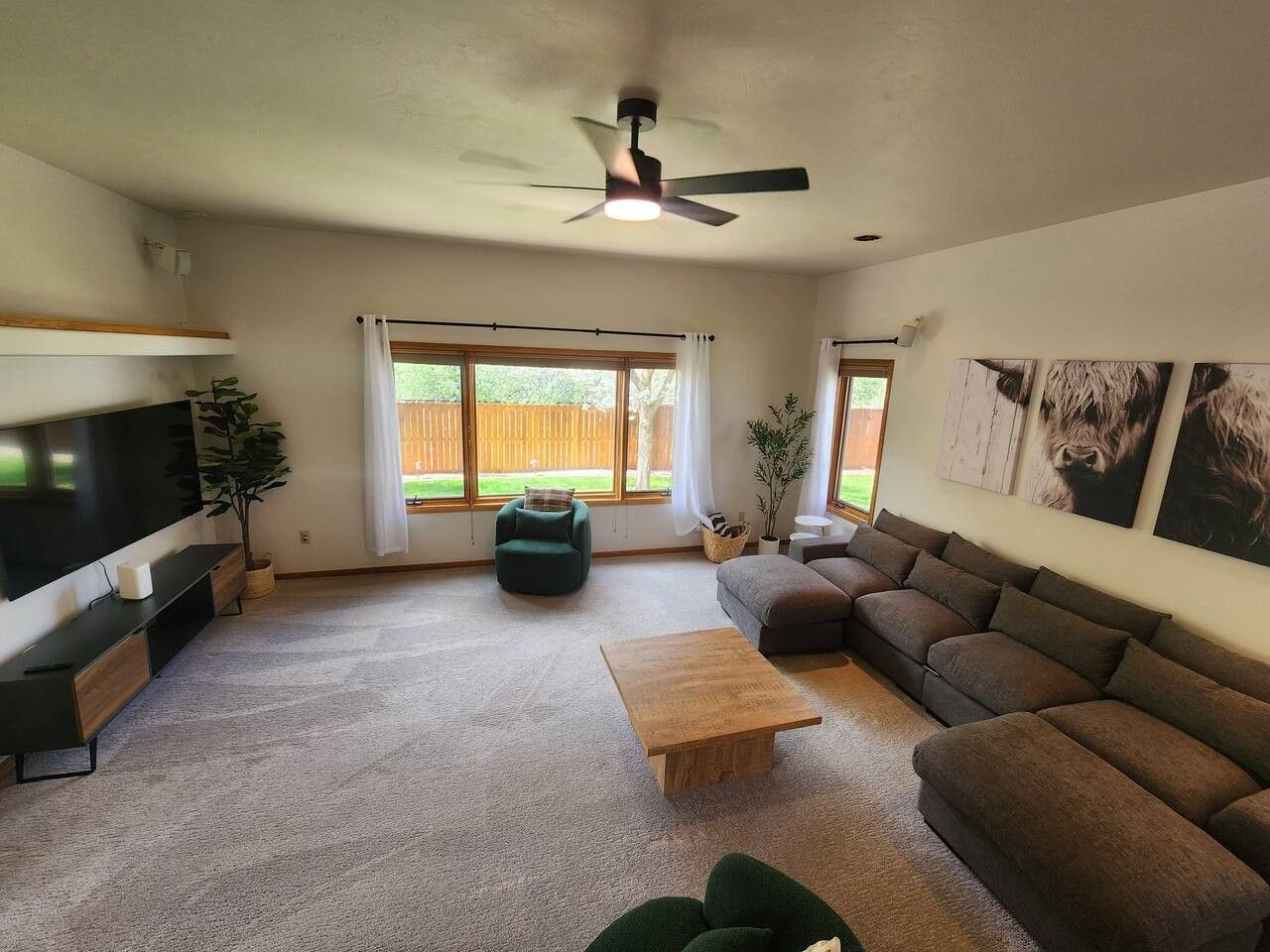 Una sala de estar con televisor de pantalla plana, ventilador de techo, sofá, mesa de centro y un ventanal con cortinas.