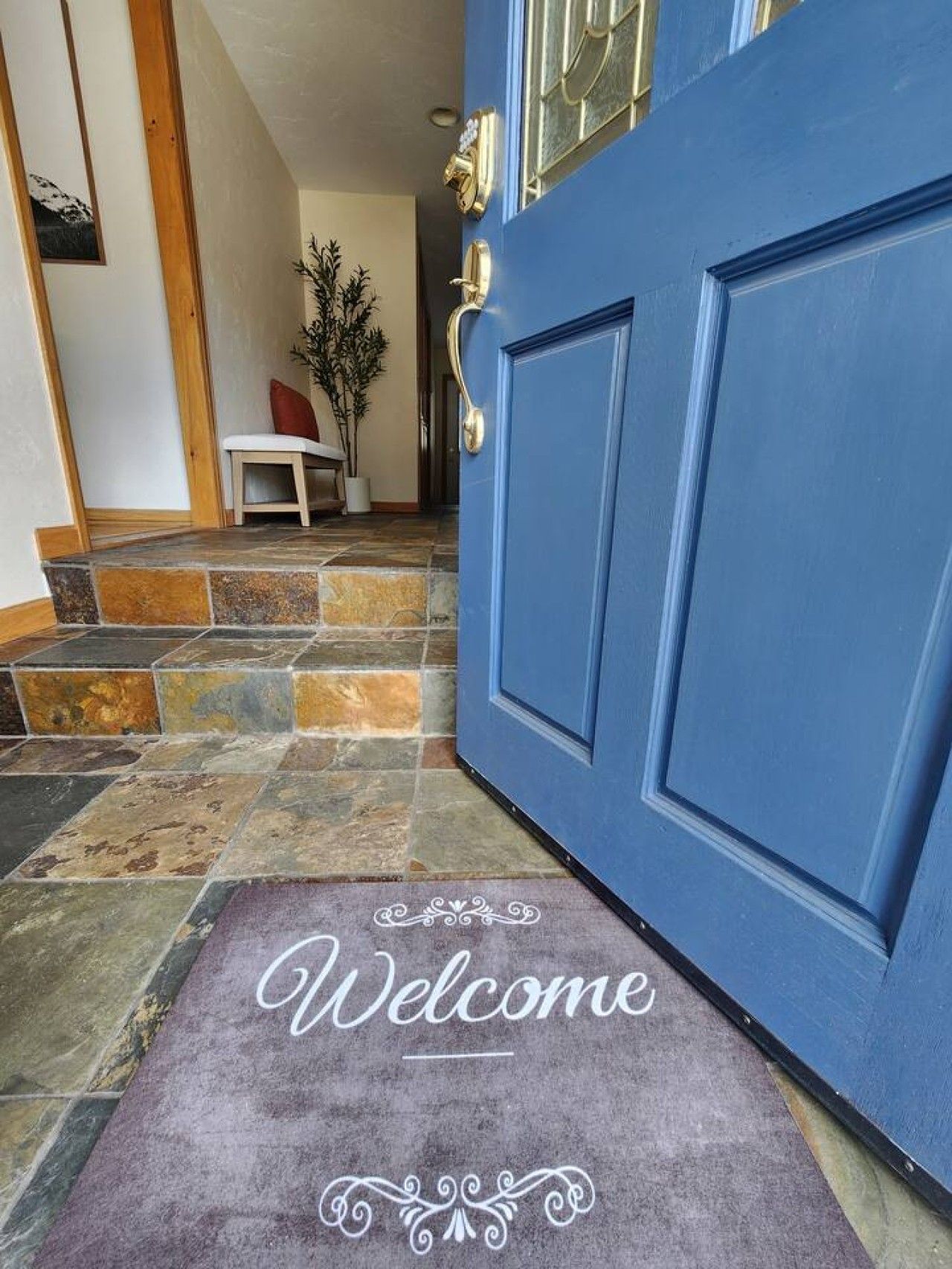 Una puerta azul con un felpudo que dice 'Welcome', una pequeña mesa con una silla y una planta en maceta.