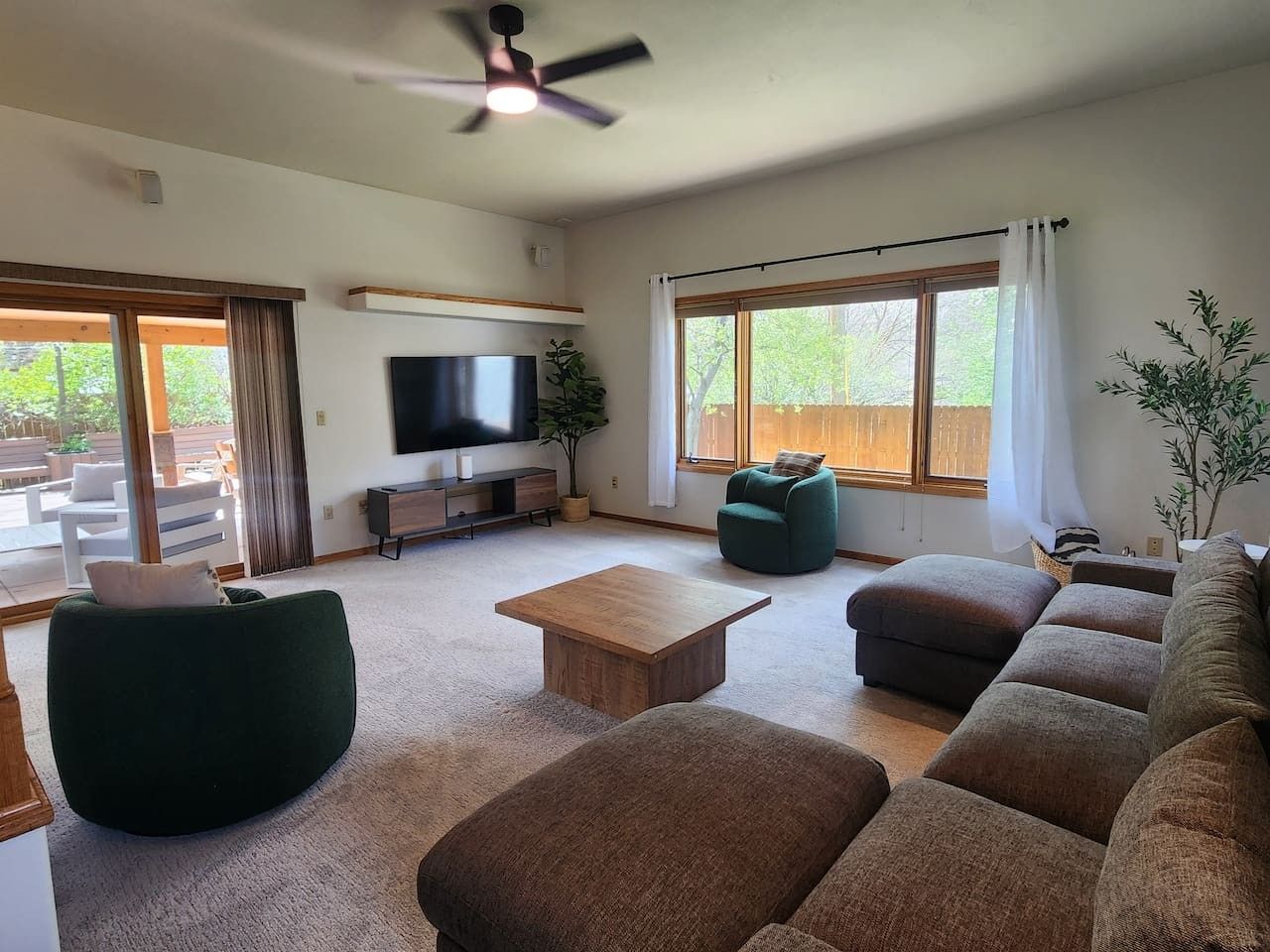 A living room with a flat-screen TV, a ceiling fan, a large window with curtains, a green armchair, a brown sofa, and a wooden coffee table.
