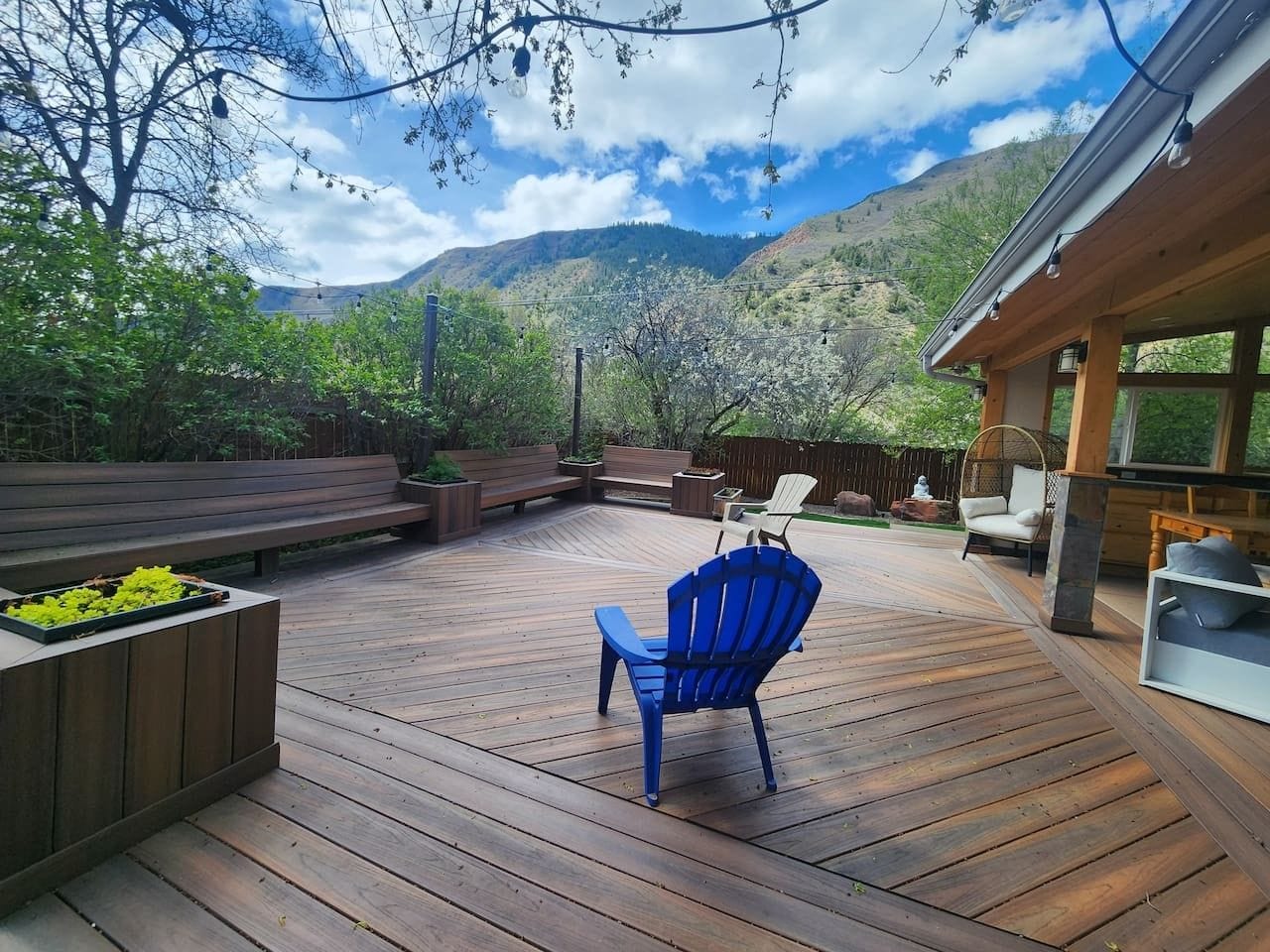 Una terraza de madera con mobiliario exterior, incluyendo una silla azul y un banco, rodeada de follaje y un paisaje montañoso.
