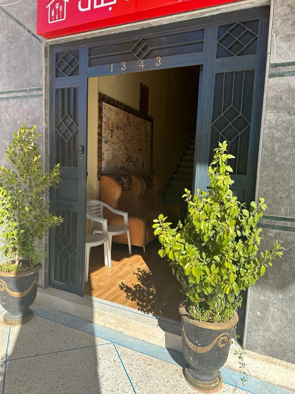 L’entrée d’un immeuble, avec le numéro « 1343 » au-dessus de la porte, un escalier à l’intérieur et deux plantes en pot à l’extérieur.