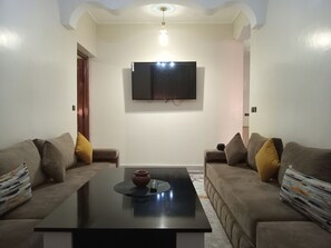 A living room with a flat-screen TV mounted on the wall, two sofas with cushions, a coffee table with a vase, and a patterned rug.