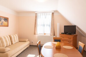 A cozy room with a sofa, a dining table with chairs, a television, and a window with curtains. - Hamr Apartmány (Nové Hamry)