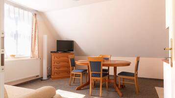 A room with a round wooden table, two chairs, a television on a dresser, a radiator, and a window with curtains.