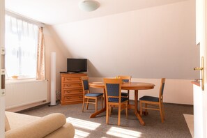 A room with a round wooden table, two chairs, a television on a dresser, a radiator, and a window with curtains. - Hamr Apartmány (Nové Hamry)