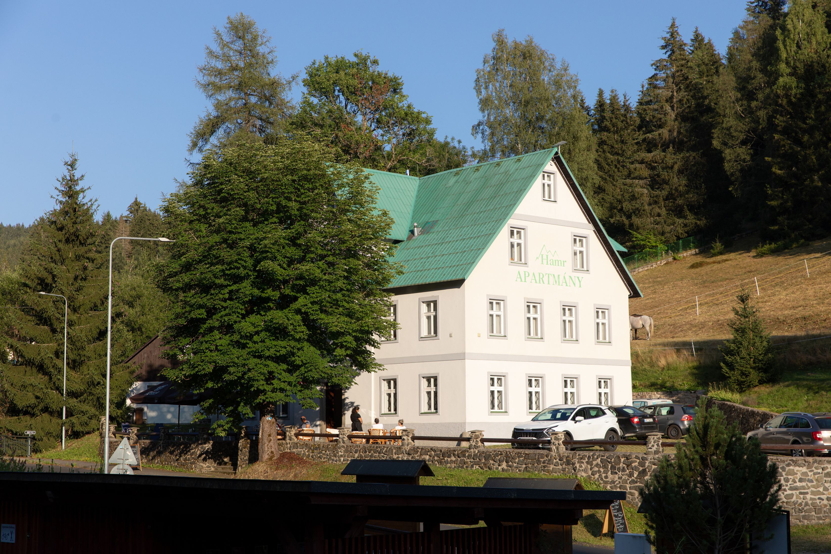A two-story building with a green roof, labeled 'Harm's APARTMENT,' surrounded by trees and a stone wall.